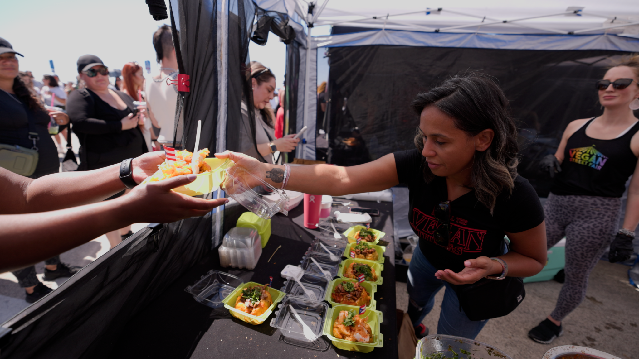 A woman wearing a black t-shirt hands a yellow container filled with vegan street food to a customer at a sunny outdoor food stall