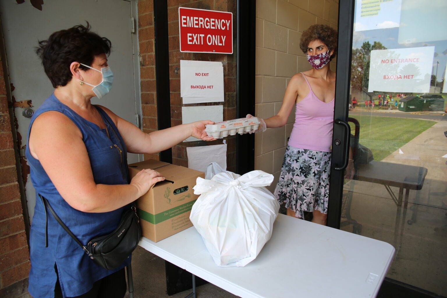 Two masked women exchanging a carton of eggs at an emergency exit doorway with grocery supplies on a table